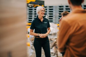 Sandrine Séjourné-Lescure, nouvelle Directrice du site U LOG de Vendargues Ambiant, souriante, portant un polo noir avec le logo U LOG, au milieu d'un entrepôt logistique de palettes.