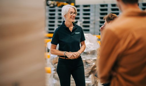 Sandrine Séjourné-Lescure, nouvelle Directrice du site U LOG de Vendargues Ambiant, souriante, portant un polo noir avec le logo U LOG, au milieu d'un entrepôt logistique de palettes.